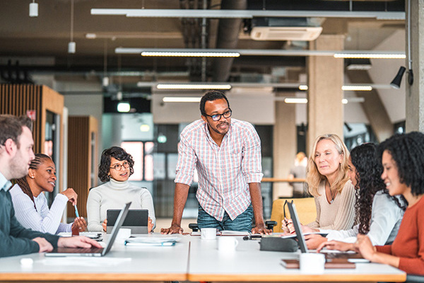 7 Diverse People Meeting At A Table With Laptops And Tablets